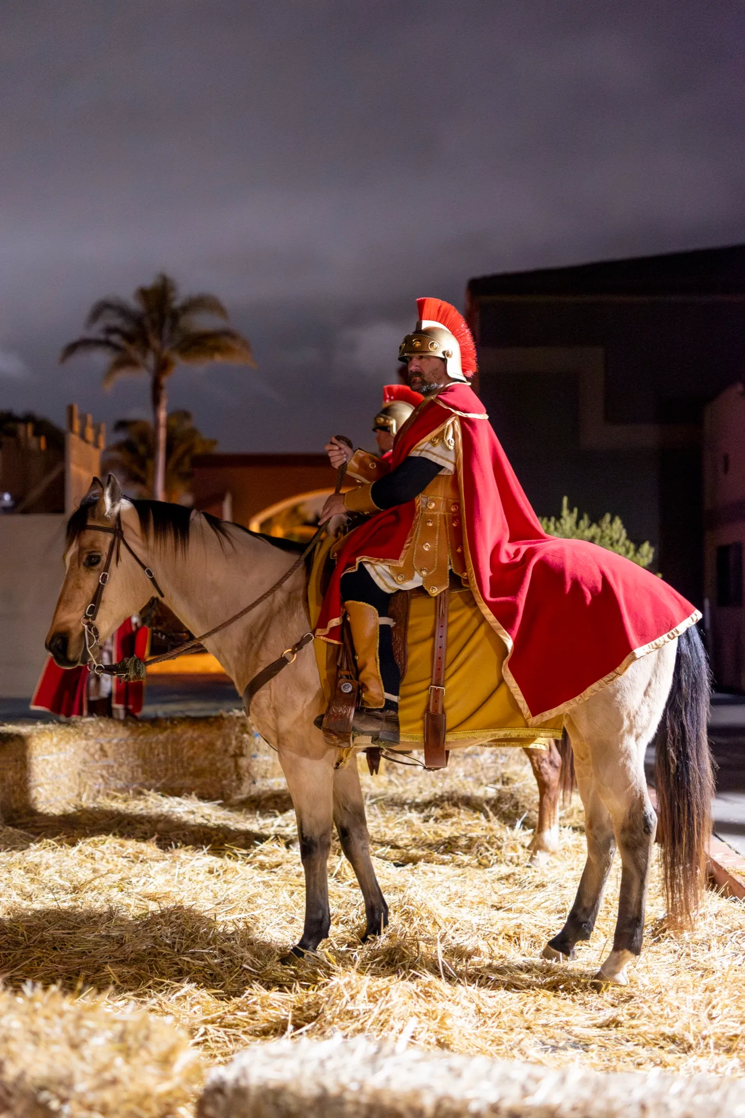 A costumed Roman soldier in a red cape rides a pale horse through straw-covered streets at night