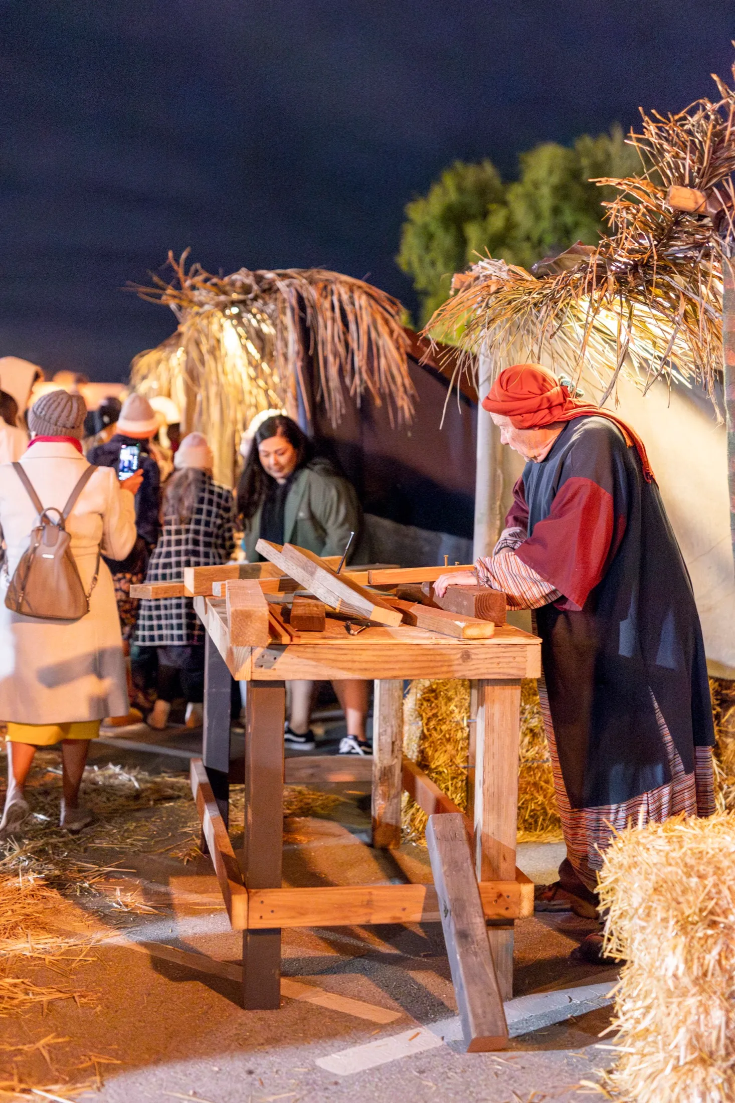 A carpenter in period costume works wood at a marketplace bench while guests gather nearby under night lights