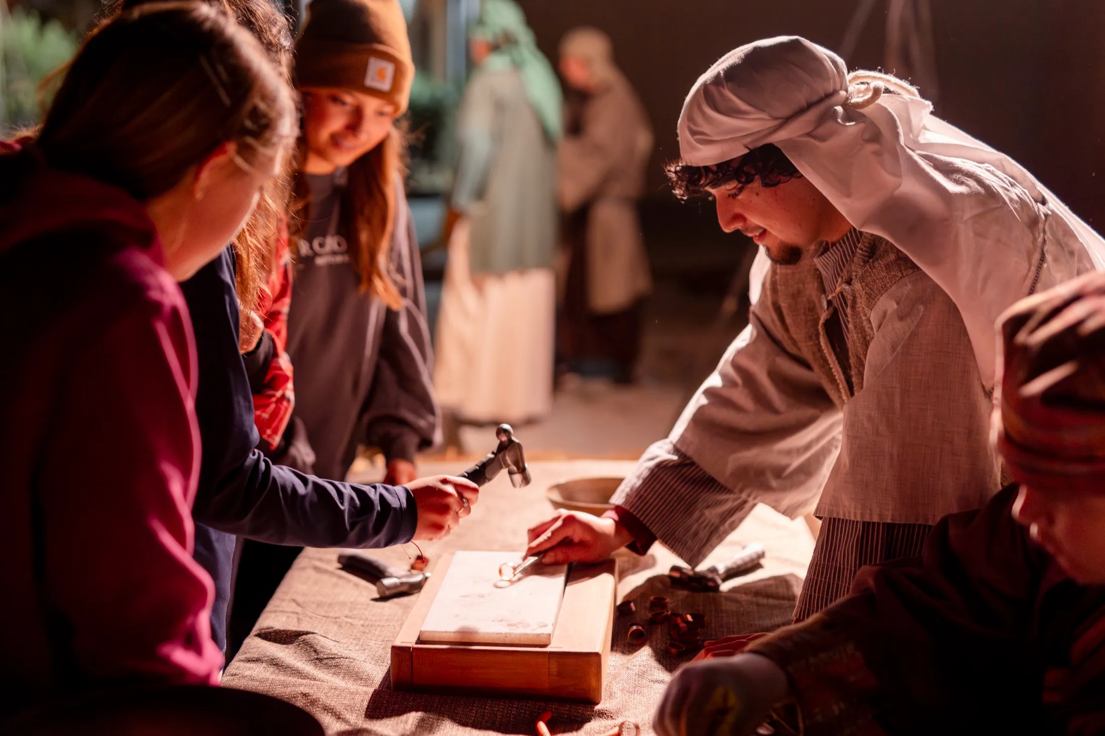 Young guests try a metalworking craft at a marketplace booth guided by a costumed volunteer