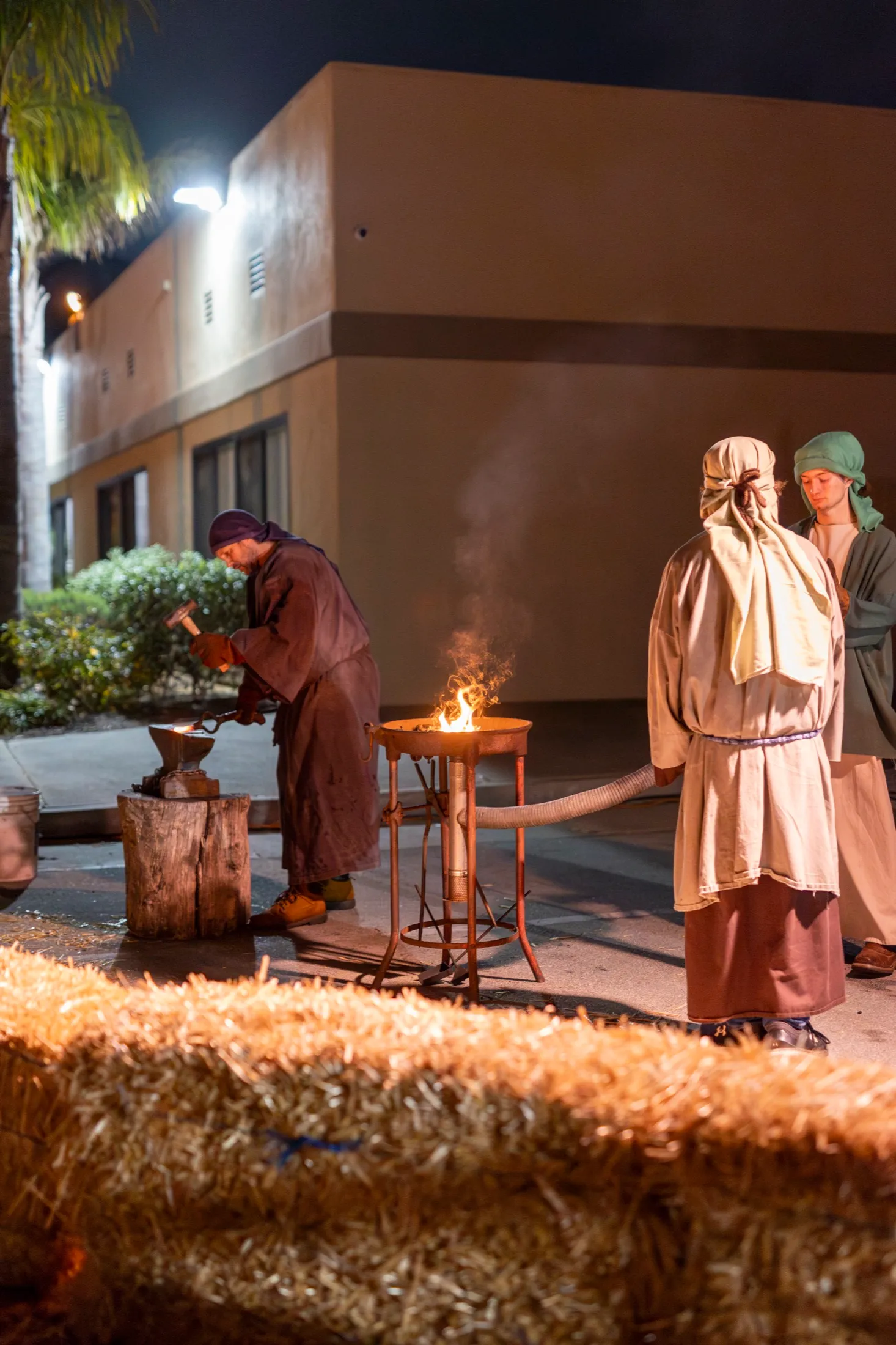A blacksmith in period costume works metal at a glowing forge while helpers work the bellows at night