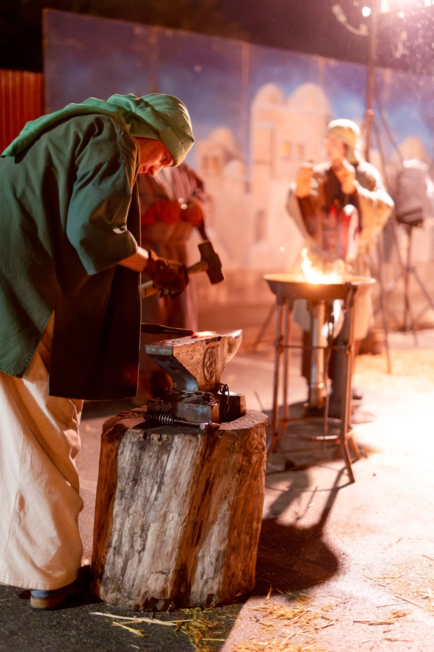 A volunteer blacksmith hammers glowing metal on an anvil set on a tree stump beside open flame