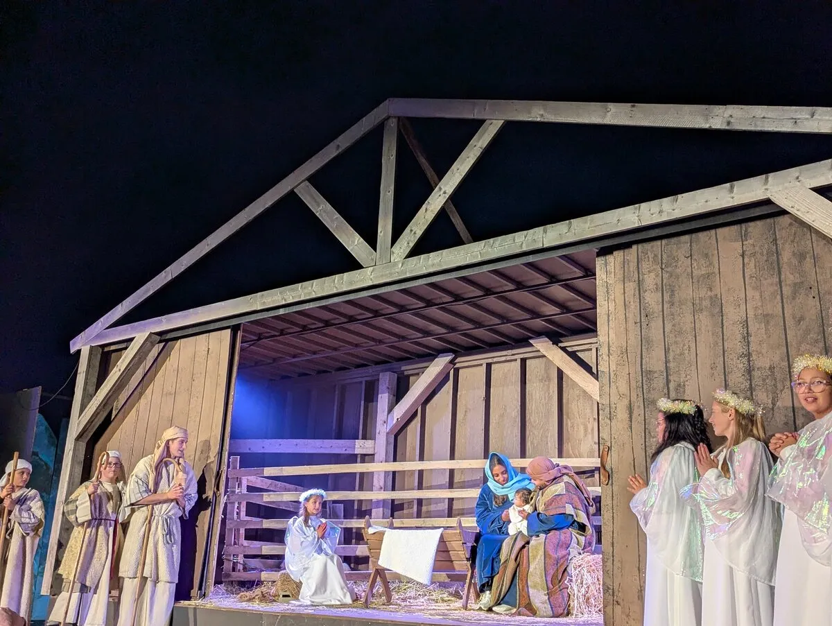 Living nativity at the manger: shepherds, Mary and Joseph with the infant, and angels under a night sky